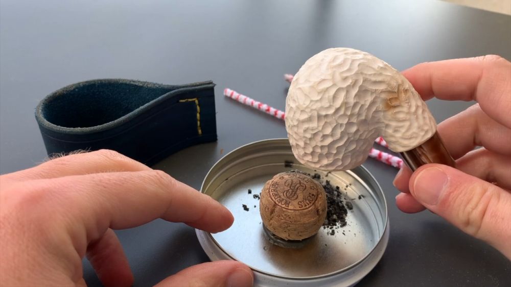 Hands holding a meerschaum pipe above a metal lid with a cork and pipe ash, demonstrating the cleaning process.